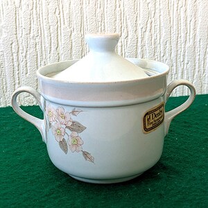 May include: A white ceramic sugar bowl with a lid, featuring a floral design in pink and gray. The bowl has two handles and a light pink band around the rim. A sticker on the side reads "Denby Fine Stoneware England."