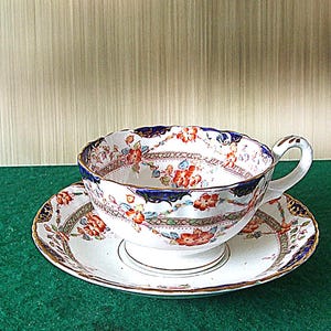 May include: A vintage teacup and saucer set. The white porcelain cup and saucer feature a floral design with red and orange flowers, blue accents, and gold trim. The cup has a curved handle and a scalloped rim.