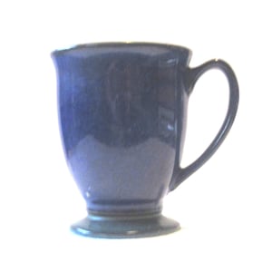 Puede incluir: Una taza de cerámica azul con asa. La taza tiene una base de pedestal y un borde ligeramente ensanchado. El color pasa de un azul más oscuro en la parte superior a un azul más claro hacia la base. La taza está sobre un fondo blanco.