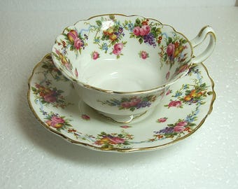 May include: A white teacup and saucer set with a floral design. The cup and saucer have a scalloped edge and gold trim. The floral pattern includes pink roses, purple grapes, and other colorful fruits.