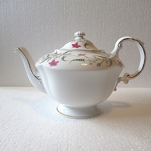 Könnte beinhalten: Weiße Porzellan-Teekanne mit floralem Design aus rosa und lila Blüten, grünen Blättern und goldenen Verzierungen. Die Teekanne hat einen geschwungenen Griff, einen Ausguss und einen Deckel mit einem kleinen Knauf. Sie steht auf einem Sockel.