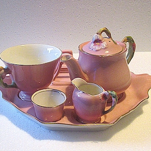 Könnte beinhalten: Ein Vintage-Teeservice aus rosa Porzellan mit Goldrand. Das Set umfasst eine Teekanne, eine Teetasse, einen Sahnekännchen und eine kleine Tasse, alles auf einem passenden Tablett. Der Teekannendeckel ist mit einer Blume verziert.