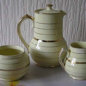 Puede incluir: Un juego de tres teteras o cafeteras de cerámica de color crema con ribetes dorados. Las teteras tienen un diseño a rayas.