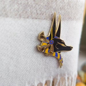 May include: Gold and black enamel pin featuring an Anubis jackal head with a blue and yellow patterned collar.