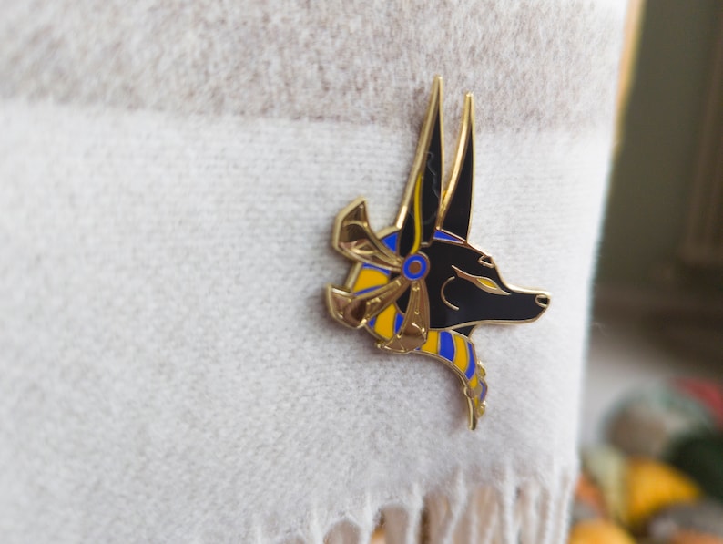 May include: Gold and black enamel pin featuring an Anubis jackal head with a blue and yellow patterned collar.