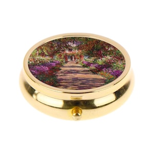 May include: A gold-colored, round pill box with a hinged lid. The lid features a colorful painting of a garden path lined with flowers. The box has a small, rounded clasp on the front.