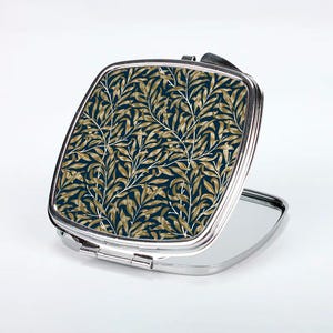 Puede incluir: Un espejo compacto plateado con un diseño floral. El diseño presenta hojas doradas sobre un fondo azul oscuro.