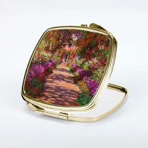 May include: A gold compact mirror with a hinged lid. The lid features a colorful painting of a garden path lined with flowers. The mirror is closed and the reflection is visible in the open side.