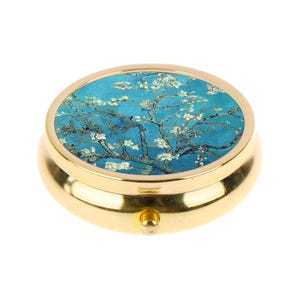 May include: A round, gold-colored pill box with a hinged lid. The lid features a painting of a tree with white blossoms against a teal background. The box has a small, gold-colored clasp.