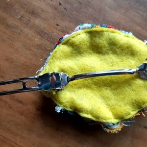 May include: A yellow felt hair clip with a colorful fabric trim and a silver metal clip.