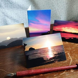 May include: Four landscape greeting cards with sunset and sunrise images. The cards show vivid colours of orange, pink, and blue. A red pen with a metal nib sits on a wooden surface, beside the cards.
