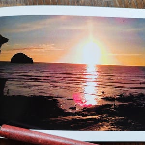 May include: A photograph of a sunset over the sea. The sun is a bright orb in the centre of the image, with its light reflecting on the water. The sky is a mix of orange, yellow, and blue. A pen is in the foreground.