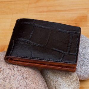 May include: A black leather wallet with a crocodile pattern and brown stitching. The wallet is folded closed and has a smooth, polished finish.