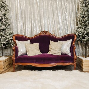 May include: A purple velvet sofa with ornate wooden carvings and three pillows, two with silver sequins and one with a beige fabric. The sofa is in front of a silver sequin backdrop with two small snow-covered Christmas trees and two nutcracker figurines on either side.
