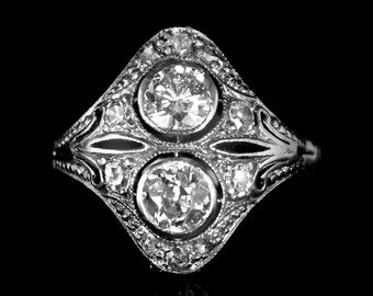 Puede incluir: Un anillo de plata con dos diamantes redondos grandes engastados en un diseño delicado y ornamentado. El anillo está rodeado de diamantes más pequeños.