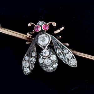 Puede incluir: Un broche de abeja de plata y oro con ojos de rubí y detalles de diamantes. La abeja está fijada en un alfiler de oro.