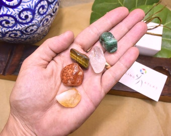 May include: A hand holding five tumbled stones: a green aventurine, a clear quartz, a tiger's eye, a carnelian, and a citrine.