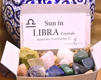 May include: A white box with a lid open, revealing a collection of crystals. The box has a label that reads "Sun in LIBRA Crystals September 23 to October 22" and a logo for Healing Atlas. The crystals are various colors, including green, pink, white, blue, and gray.
