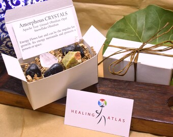May include: A white box with a brown paper filler and a variety of crystals. The box is open and the crystals are visible. The box has a label that reads "Amorphous Crystals" and lists the types of crystals included: Apache Tear, Girasol Obsidian, Opal, and Snowflake Obsidian. The label also includes a description of the crystals' energy properties. The box is sitting on a wooden surface with a business card that reads "Healing Atlas".