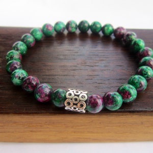 May include: A beaded bracelet featuring green and maroon gemstones. The bracelet is strung with round beads and has a silver-colored cylindrical accent piece. The gemstones have a speckled appearance, and the bracelet is displayed on a wooden surface.