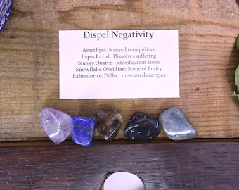 May include: A set of six tumbled stones, including amethyst, lapis lazuli, smoky quartz, snowflake obsidian, and labradorite. The stones are arranged on a wooden surface with a card that reads "Dispel Negativity" and lists the properties of each stone.
