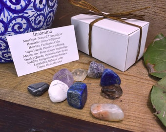 May include: A collection of tumbled gemstones, including amethyst, lapis lazuli, and smoky quartz, arranged on a wooden surface. A white gift box tied with a brown ribbon sits nearby, along with a card listing the stones' properties. The card reads "Insomnia".
