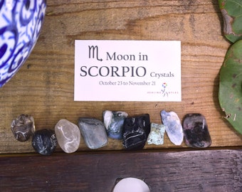 May include: A white card with the text "♏ Moon in SCORPIO Crystals October 23 to November 21" and a colorful logo for Healing Atlas. A row of 10 small, colorful crystals on a wooden surface.