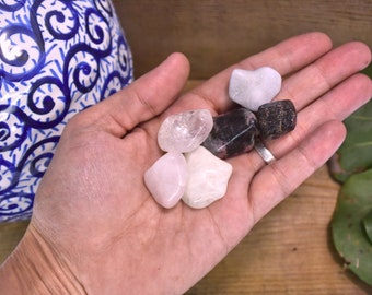 May include: A collection of six tumbled heart-shaped stones in various colors, including white, pink, and black. The stones are arranged in a hand.