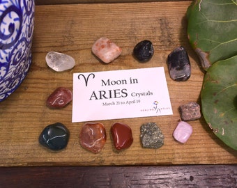 May include: A set of crystals for the zodiac sign Aries. The crystals are arranged in a circle around a card that reads "Moon in Aries Crystals March 21 to April 19". The crystals are various colors, including red, green, black, and pink.