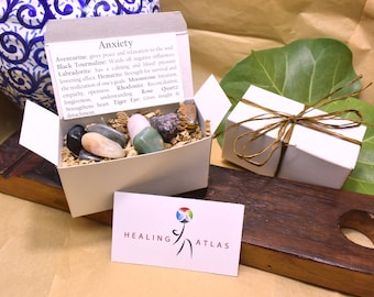 May include: A white box filled with various crystals and stones, including aventurine, black tourmaline, labradorite, hematite, moonstone, rhodonite, rose quartz, and tiger's eye. The box is labeled "Anxiety" and includes a description of the healing properties of each stone. The box is sitting on a wooden surface with a business card that reads "Healing Atlas".