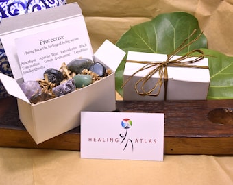 May include: A white box filled with a variety of crystals, including amethyst, apache tear, labradorite, black tourmaline, green aventurine, lepidolite, and smoky quartz. The box is labeled "Protective: bring back the feeling of being secure". A business card for Healing Atlas is in the foreground.