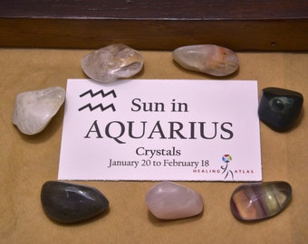 May include: A card with the astrological sign Aquarius and the text "Sun in Aquarius Crystals January 20 to February 18" surrounded by various crystals, including clear, pink, black, and multi-colored.