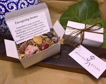 May include: A white box labeled "Energizing Stones" contains various colorful gemstones. The stones include carnelian, citrine, red jasper, garnet, ruby, and sunstone. A second white box is tied with brown twine. A tag reads "Healing Atlas."