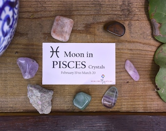 May include: A white card with the text "Moon in PISCES Crystals February 19 to March 20" and the Pisces astrological symbol.  There are several crystals on a wooden surface, including amethyst, rose quartz, and green aventurine.