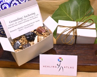 May include: A white box filled with six grounding crystals: hematite, obsidian, petrified wood, smoky quartz, and picture jasper. The box is open and the crystals are visible. The box is labeled "Grounding/Anchoring" and includes a description of the crystals. The box is sitting on a wooden surface. A business card for "Healing Atlas" is in the foreground.