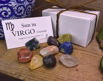 May include: A white box with a brown ribbon tied around it. The box contains a set of crystals for the Virgo zodiac sign. The crystals are arranged on a wooden surface. The box is labeled "Sun in Virgo Crystals August 23 to September 22".