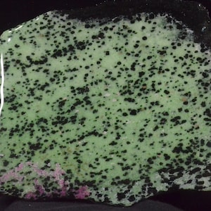 May include: A green and black speckled stone with a smooth, polished surface. The stone has a mottled appearance with dark green and black spots.