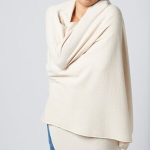 May include: A person wearing a cream-colored knitted scarf wrapped around their shoulders. The scarf is draped over the person's arms and is made of a soft, cozy material.