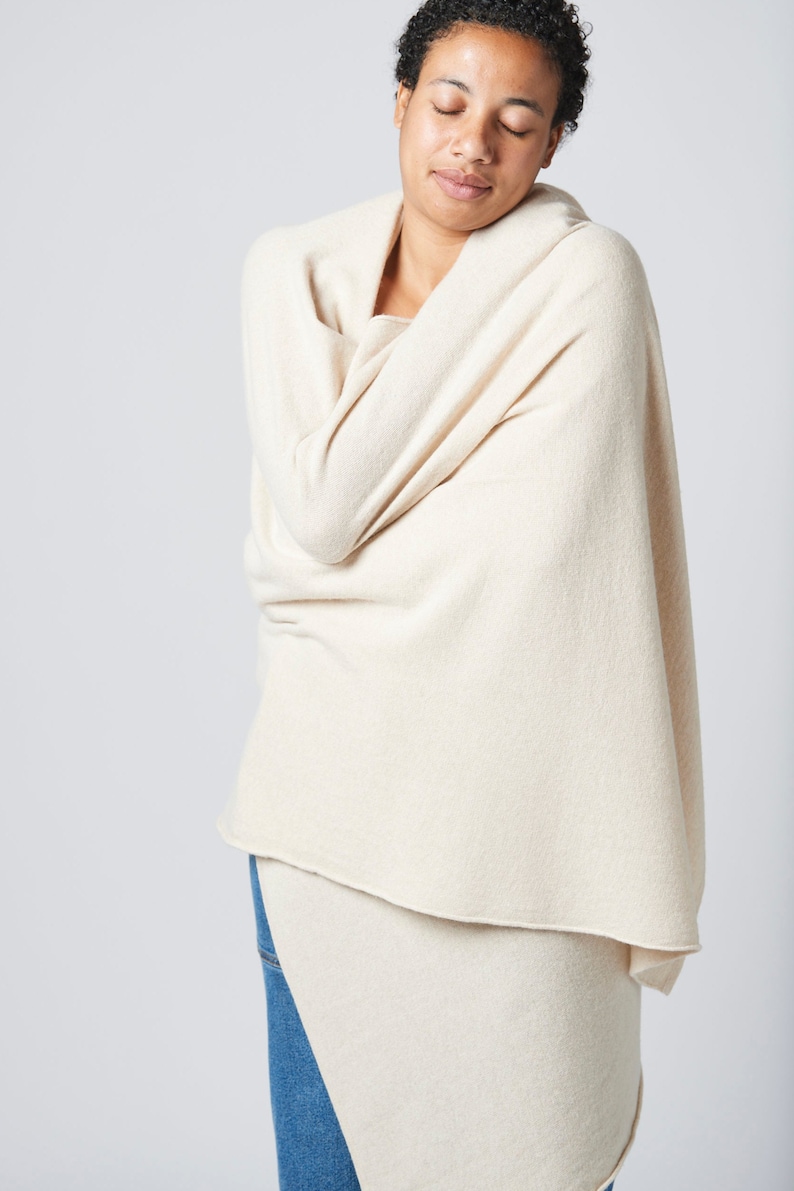 May include: A person wearing a cream-colored knitted scarf wrapped around their shoulders. The scarf is draped over the person's arms and is made of a soft, cozy material.