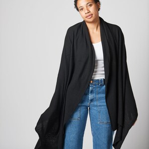May include: A black knit wrap with a long, flowing design. The wrap is draped over a person wearing a white tank top and blue jeans.