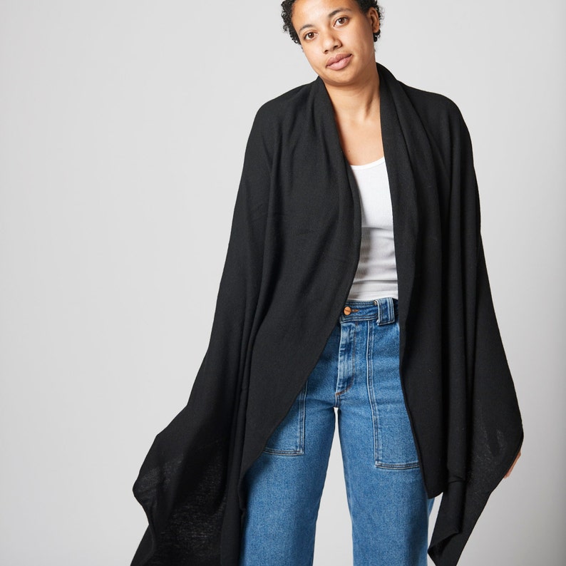 May include: A black knit wrap with a long, flowing design. The wrap is draped over a person wearing a white tank top and blue jeans.