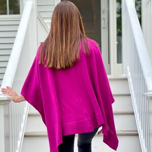 May include: A woman wearing a magenta knitted poncho with a long, straight hairstyle. The poncho has a loose fit and a slightly asymmetrical hemline.