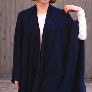 May include: A navy blue knitted wrap with a long, flowing design. The wrap is draped over a person's shoulders and is worn with a white shirt.