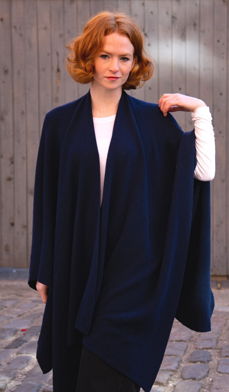May include: A navy blue knitted wrap with a long, flowing design. The wrap is draped over a person's shoulders and is worn with a white shirt.