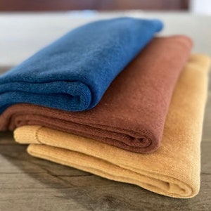 May include: Three folded fabric blankets in different colors: a dark blue, a rust brown, and a light yellow.
