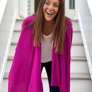 May include: A woman wearing a bright pink knitted cardigan over a white t-shirt and black pants. She is standing on a set of white stairs and smiling.