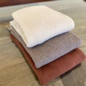 May include: A stack of three sweaters in different colors: beige, brown, and rust. The sweaters are folded and arranged on a wooden surface.