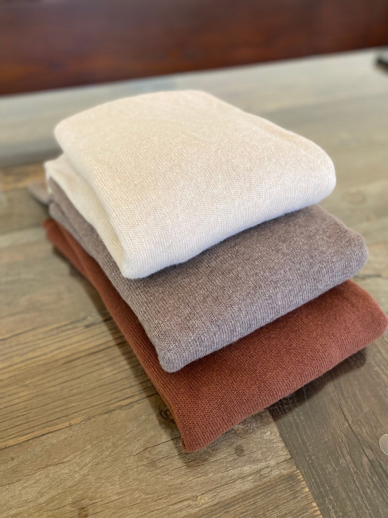 May include: A stack of three sweaters in different colors: beige, brown, and rust. The sweaters are folded and arranged on a wooden surface.