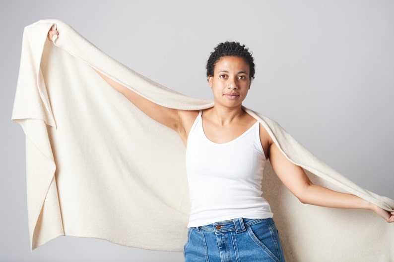 May include: A person wearing a white tank top and blue jeans is holding a large, off-white blanket in front of them.