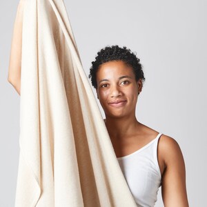 May include: A person holds up a large, off-white towel. The towel is soft and fluffy, and it appears to be made of a natural material. The person is smiling and looking at the camera.